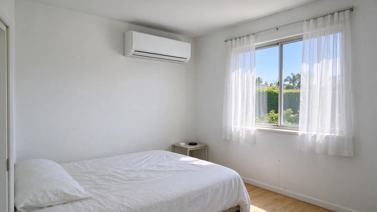 Upstairs bedroom in a San Diego house on a bright summer day, curtains half drawn over a sunny window and a white wall-mounted mini split head high on the solid wall beside it